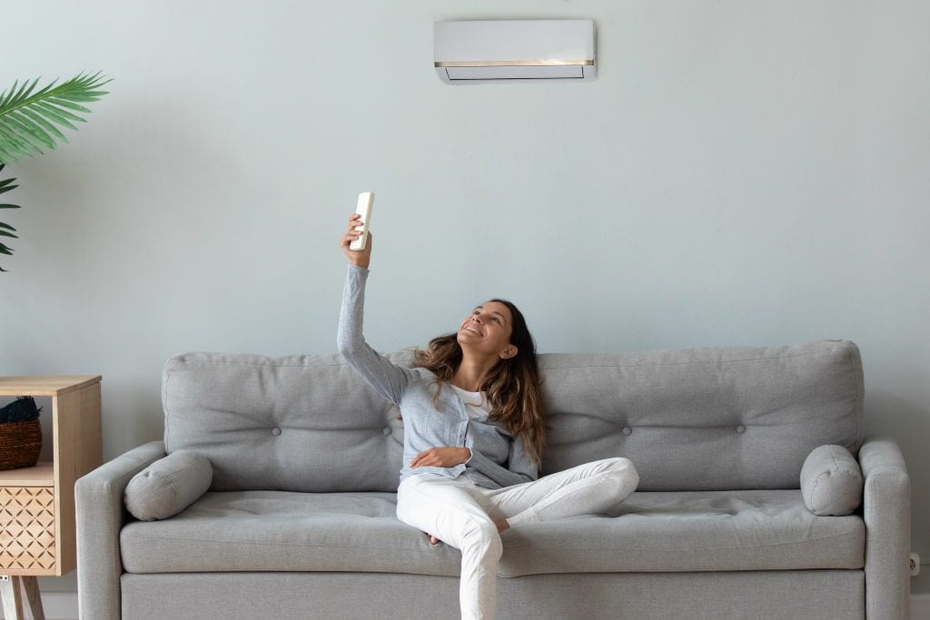 Smiling young woman relaxing on couch in living room, using air conditioner remote controller, switching, beautiful girl setting comfort temperature at home, enjoying fresh air, resting on sofa